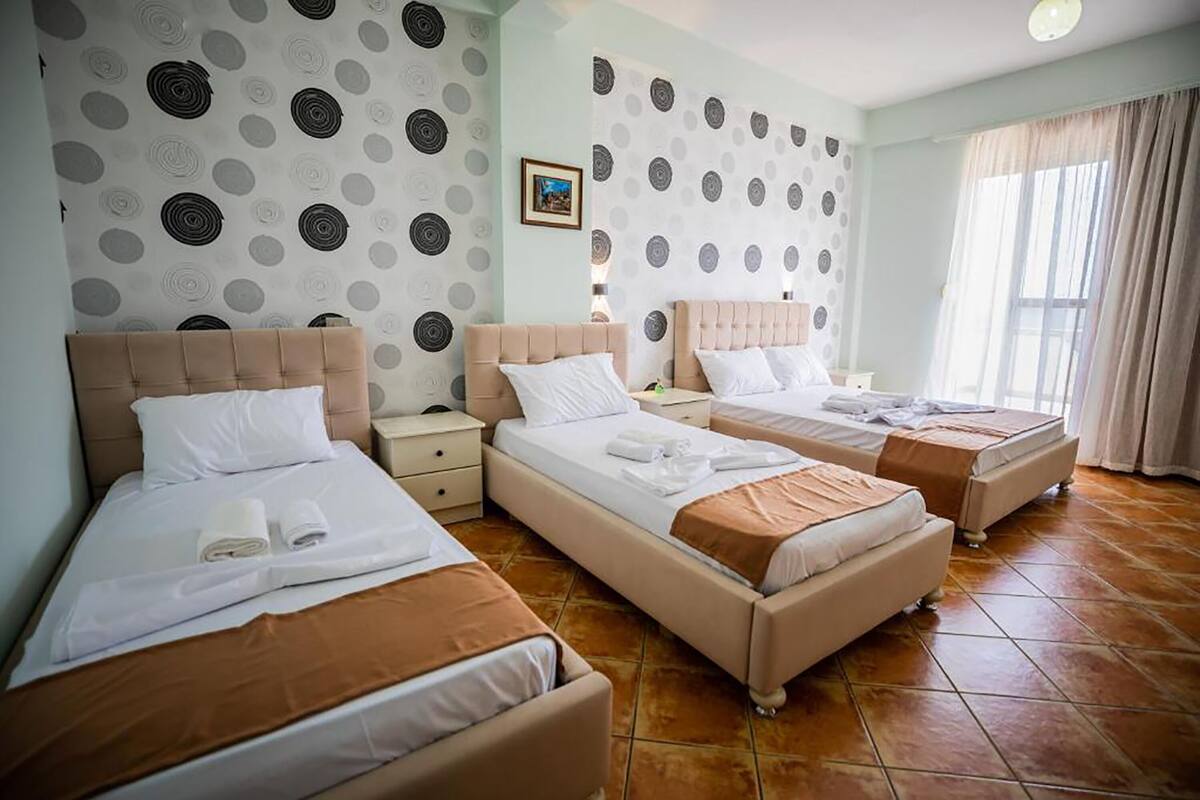 A hotel room with three beds, each with a headboard and pillows. There is a small bedside table with a lamp on each bed. The room has a tiled floor and a window with curtains.