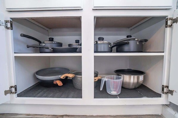 A kitchen cabinet with four shelves, each holding various kitchenware items like pots, pans, and a measuring cup.