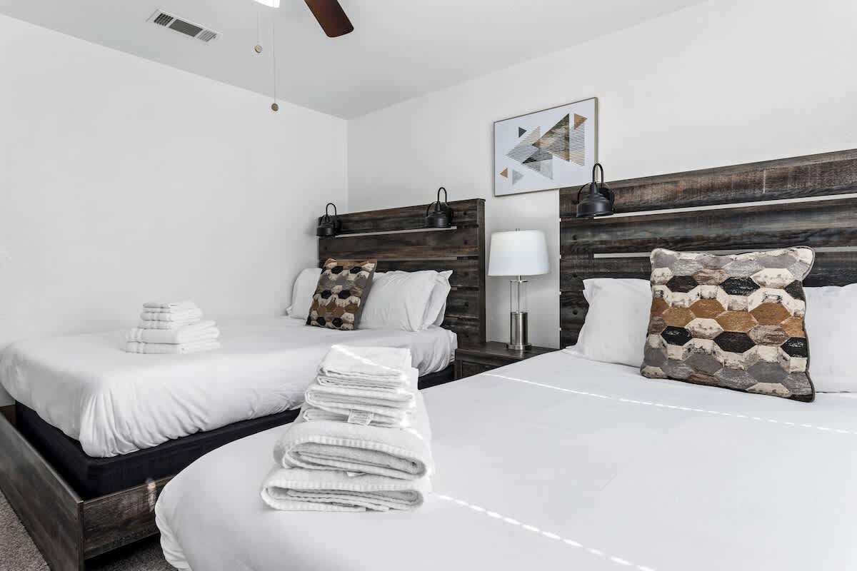 Une chambre d’hôtel avec deux lits, chacun agrémenté de draps blancs et de coussins décoratifs. On y trouve une tête de lit en bois, une lampe fixée au mur et un tableau encadré au-dessus des lits.