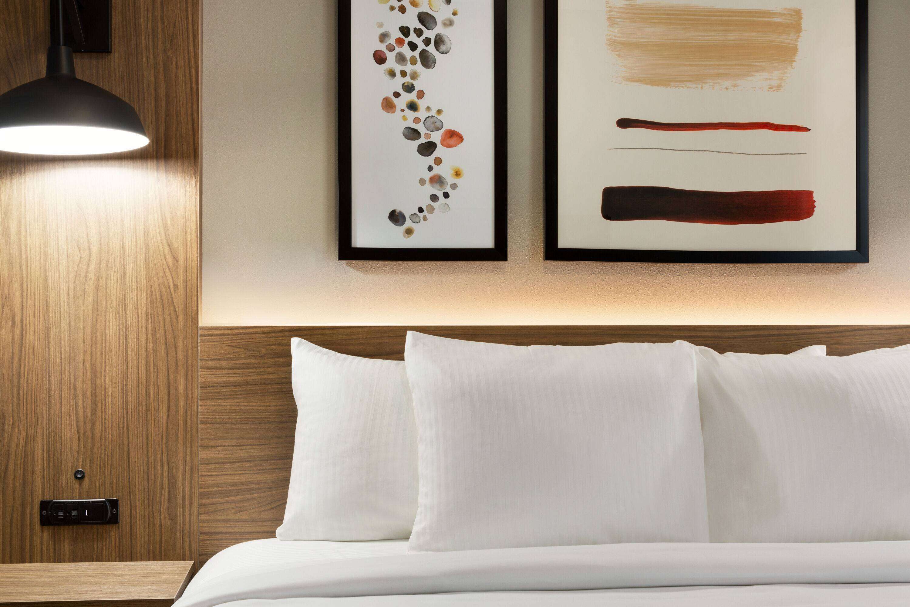 A neatly made bed with white linens, a wooden headboard, and two framed abstract paintings on the wall above.