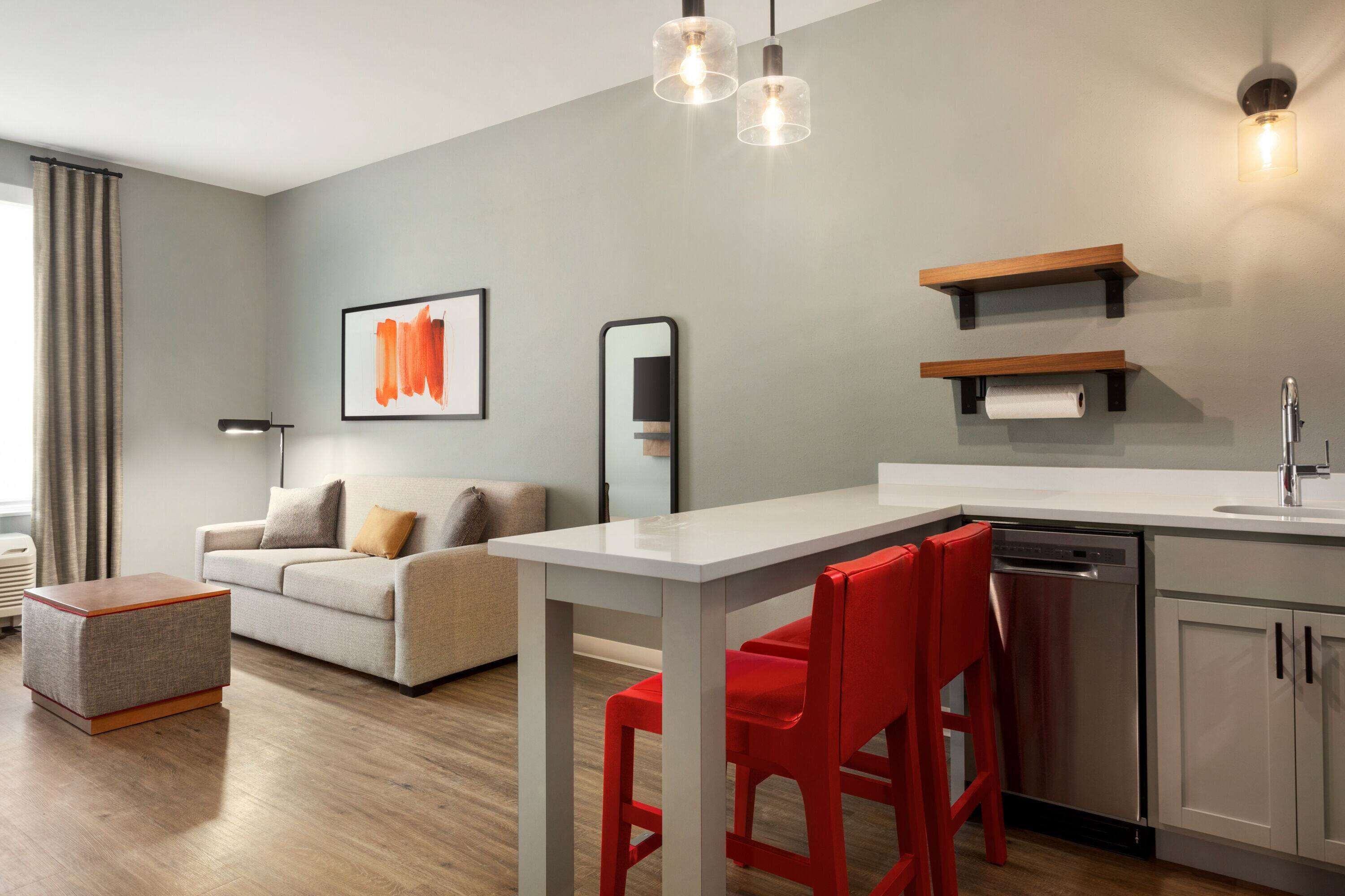 A modern living room with a sofa, a dining table with red chairs, a kitchen area with a dishwasher, and a large mirror on the wall.