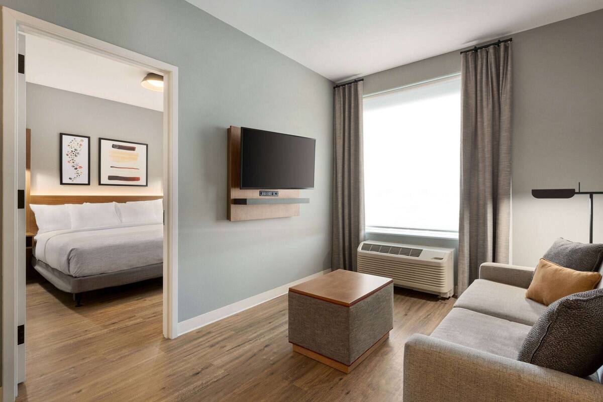 Una habitación de hotel moderna con cama, sofá, televisión y ventana con cortinas.