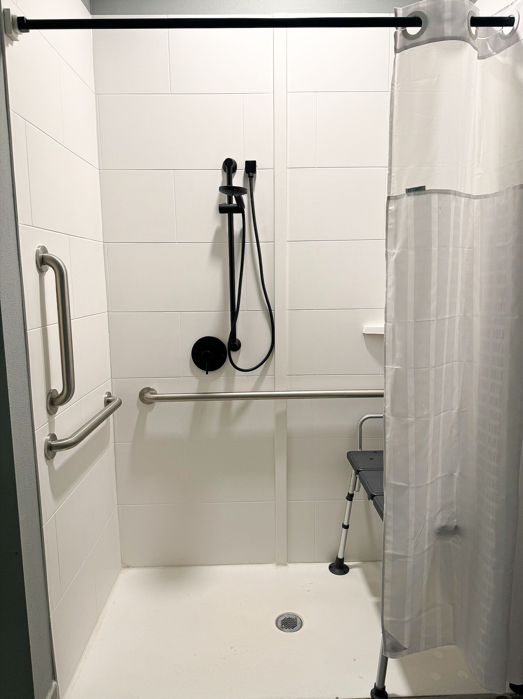 Un box de ducha con paredes de azulejos blancos, una barra de apoyo, un cabezal de ducha manual y una cortina de ducha.
