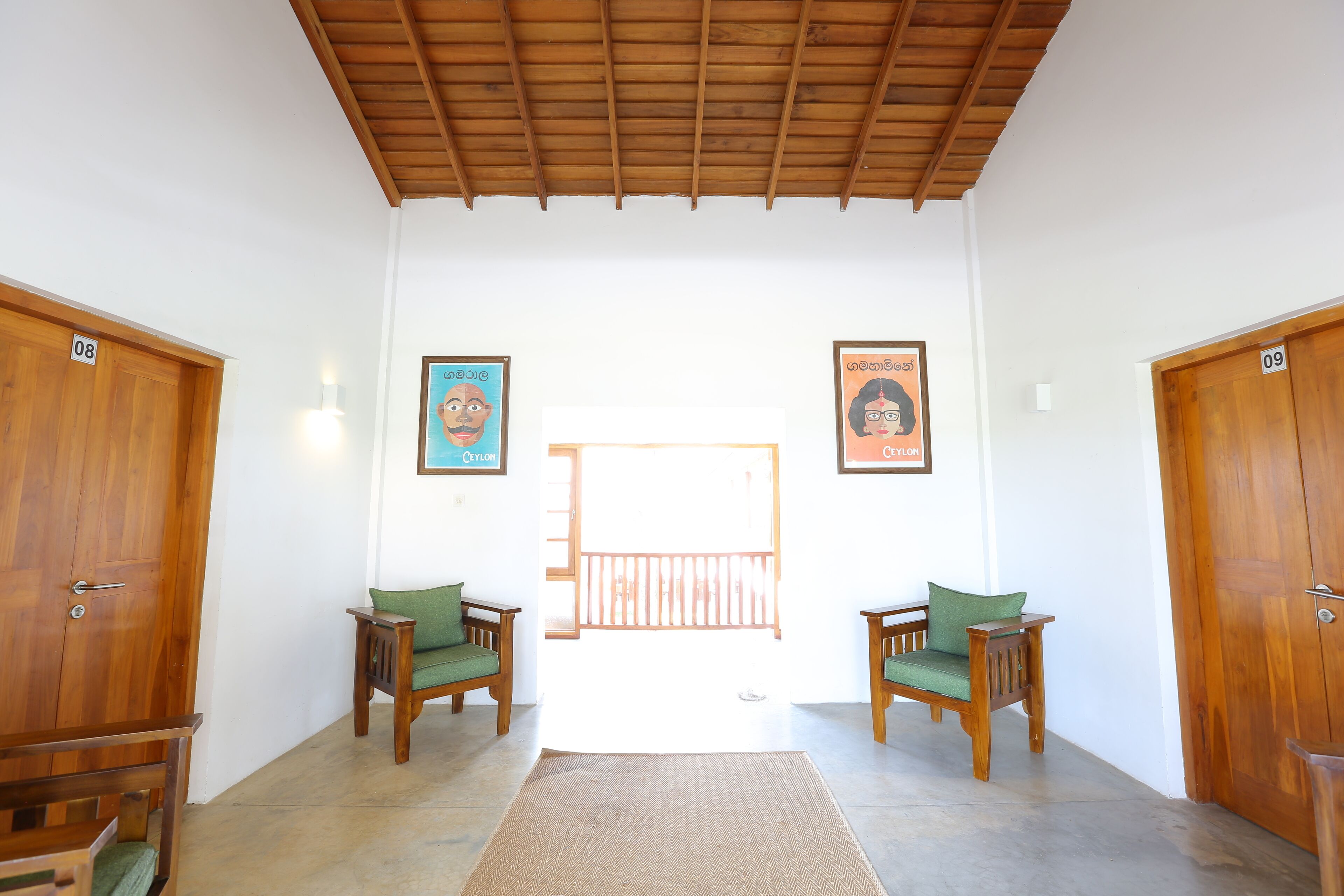 Un couloir avec des portes en bois numérotées 08 et 09, deux chaises en bois avec des coussins verts, et un grand tapis tissé sur le sol.