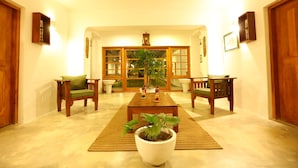 A living room with a wooden coffee table, two green armchairs, a large potted plant, and a wooden cabinet with a picture.