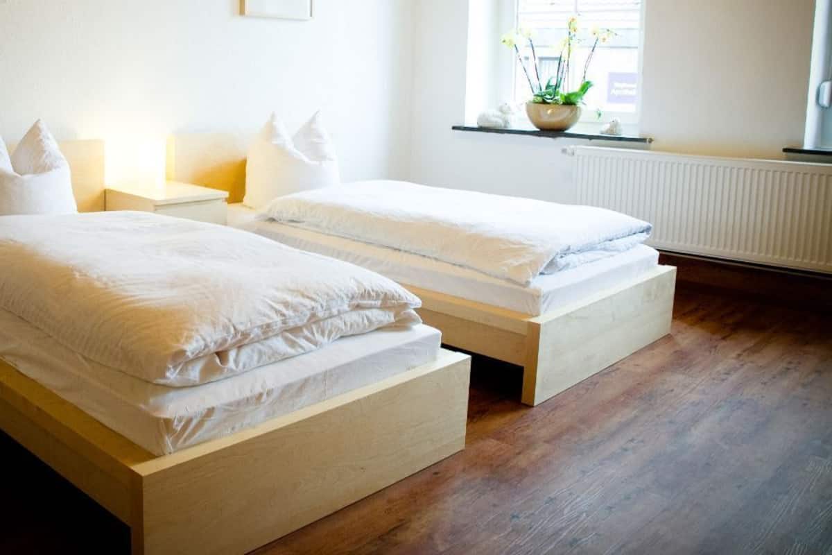 A bedroom with two single beds, wooden headboards, white bedding, bedside tables with lamps, a window with a view of the outside, and a radiator.