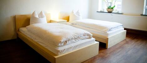 Un dormitorio con dos camas individuales, cabeceros de madera, ropa de cama blanca, mesitas de noche con lámparas, una ventana con vistas al exterior y un radiador.
