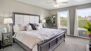 Une chambre avec un grand lit, un ventilateur de plafond, une fenêtre avec des rideaux et une plante.