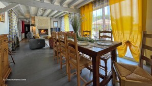 A dining area with wooden chairs and a table set for four, a fireplace, and a view through large windows with yellow curtains.