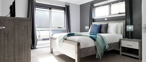 Un dormitorio con cama, mesitas de noche, un armario y ventanas amplias.