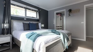 A bedroom with a bed, bedside tables, a window with curtains, and a door leading to another room. - Happy Tails Devonport (East Devonport)