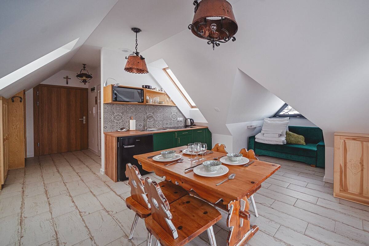 Cozinha compacta com micro-ondas, pia e armários verdes. Há uma mesa de jantar posta com pratos e copos. Um sofá verde e um armário de madeira estão na área de estar.