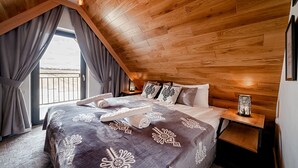 A bedroom with a sloped ceiling, a bed with patterned bedding, two bedside tables with lamps, and a view of the beach through the window.