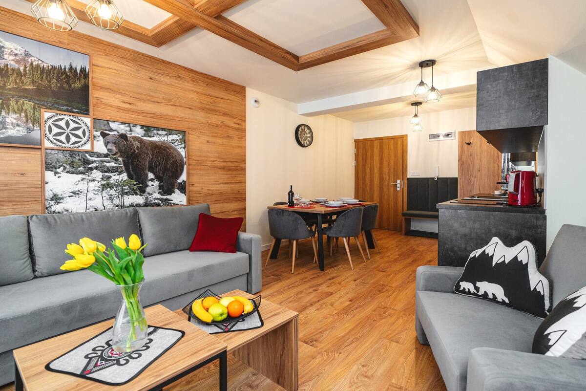 Uma sala de estar moderna com um sofá cinza, uma mesa de centro com um vaso de flores amarelas, e uma área de jantar com uma mesa e cadeiras de madeira.