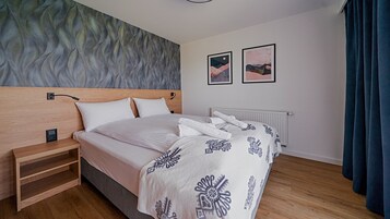 Un dormitorio moderno con cabecera de madera, cama con ropa de cama blanca y mantas estampadas, mesita de noche, lámpara de pared y dos cuadros enmarcados.