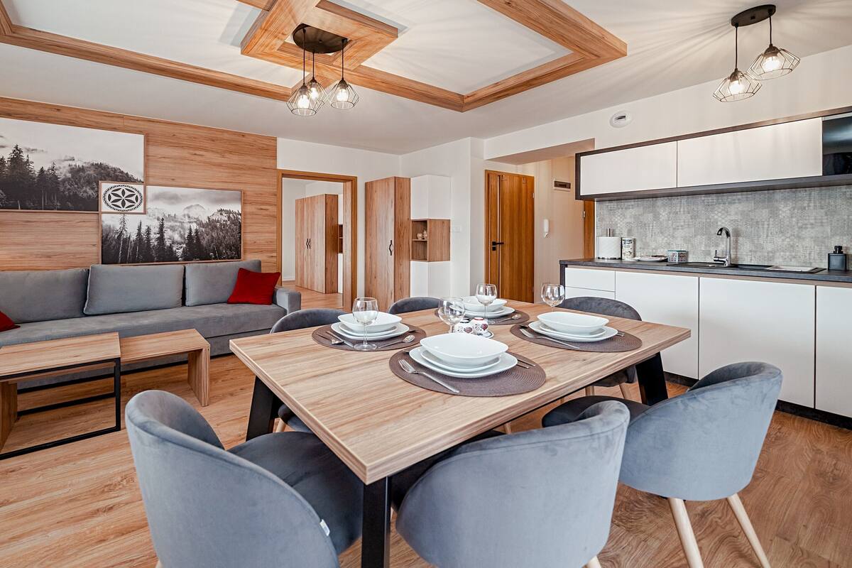 Cozinha moderna com fogão embutido, área de jantar com mesa de madeira e cadeiras cinzentas, e sala de estar com sofá cinza e almofadas vermelhas.