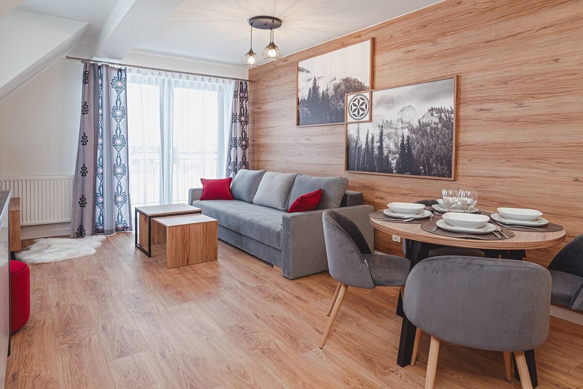 Uma sala de estar moderna com um sofá cinza, uma mesa de jantar de madeira posta para quatro pessoas e uma grande janela com cortinas estampadas.