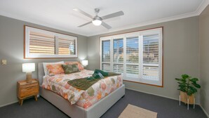 A bedroom with a bed, a ceiling fan, a nightstand, a lamp, a window with blinds, and a potted plant. - Akuli By the Sea - Unit 1 - FNHolidays (Iluka)
