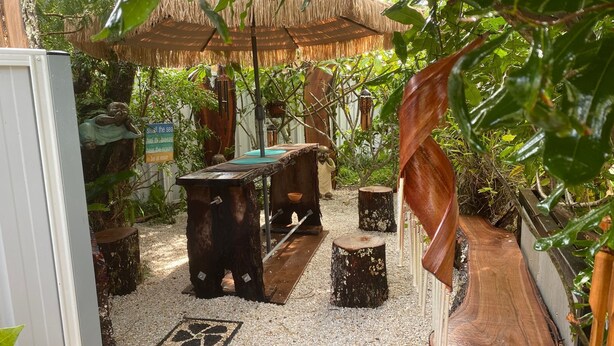 A rustic outdoor seating area with a wooden table and benches, surrounded by greenery and a thatched roof structure. - Akuli By the Sea - Unit 1 - FNHolidays (Iluka)