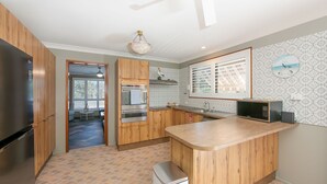 A modern kitchen with wooden cabinets, a built-in oven, a microwave, and a large window with blinds. - Akuli By the Sea - Unit 1 - FNHolidays (Iluka)