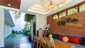 Un interior modern amb una taula de dinar de fusta, cadires de vimet, un sofà amb coixins, una paret de maó amb vegetació i vista a una piscina i zona exterior.