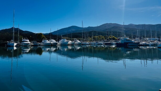A marina with boats docked, calm water, and mountains in the background.