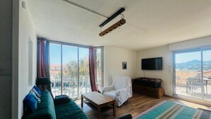 A room with a green sofa, a wooden table, a white chair, a TV, and a balcony with a view.