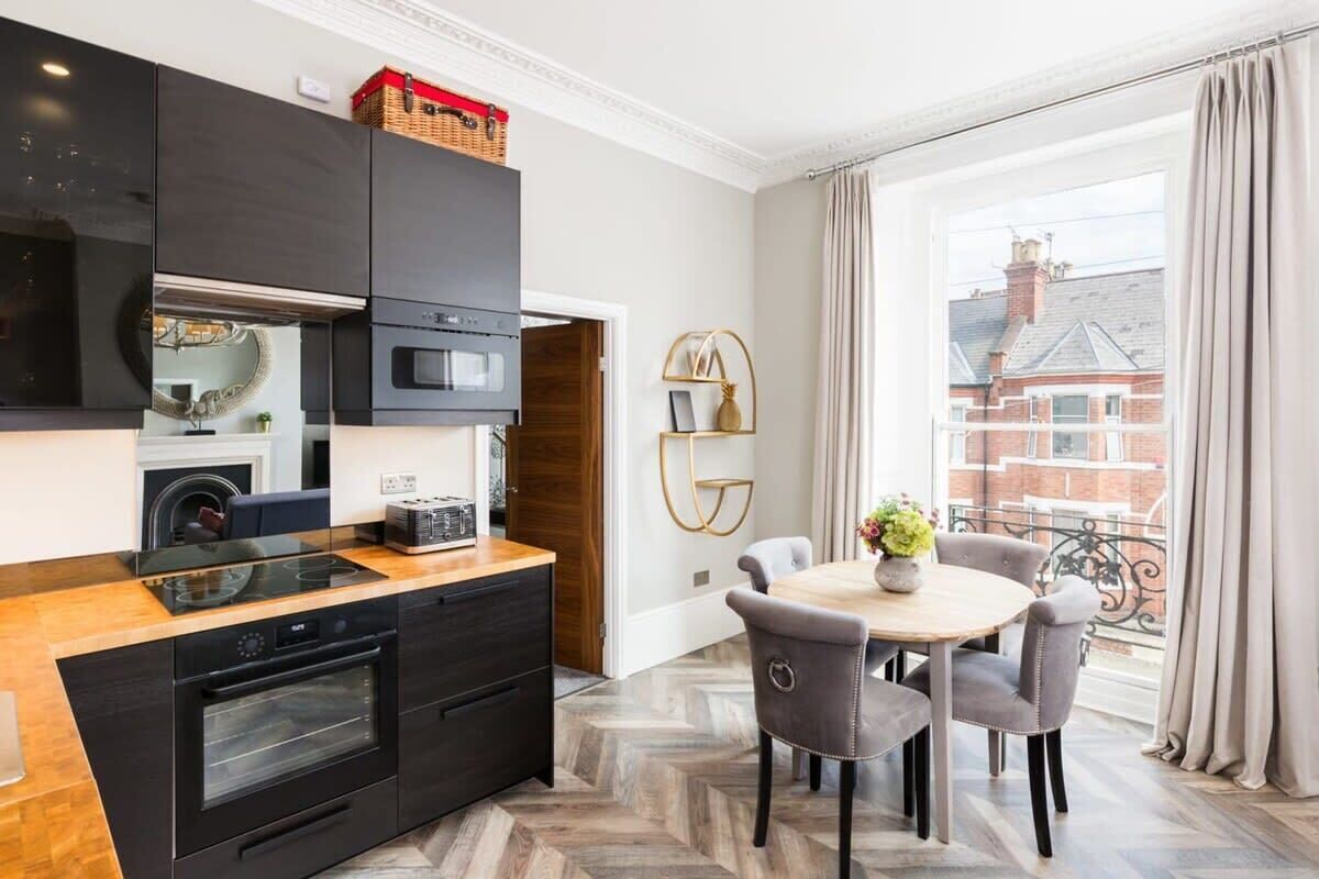 A modern kitchen with black cabinets, a wooden countertop, and a built-in oven. There is a dining area with a round table and chairs, and a view of a balcony with curtains.