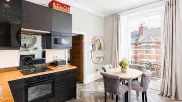 A modern kitchen with black cabinets, a wooden countertop, and a built-in oven. There is a dining area with a round table and chairs, and a view of a balcony with curtains.