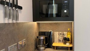 A compact kitchen with a microwave, coffee maker, and a small wooden shelf with kitchen essentials. - Ground floor compact studio with mini kitchen (Warrington)