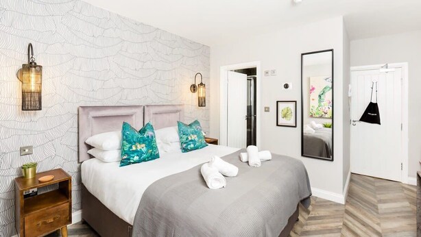 A hotel room with a large bed, two wall sconces, a mirror, and a door leading to another room. - Ground floor compact studio with mini kitchen (Warrington)