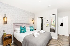 A hotel room with a large bed, two wall sconces, a mirror, and a door leading to another room. - Ground floor compact studio with mini kitchen (Warrington)