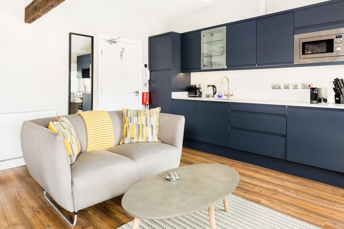 Un salon moderne avec un canapé gris, une table basse et un coin cuisine doté de meubles sombres et d’un four à micro-ondes.