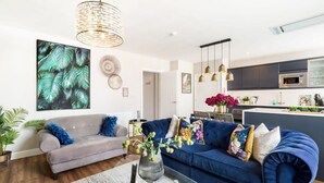 Ein modernes Wohnzimmer mit einem grauen Sofa, einem blauen Sofa und einem Glastisch mit einer Blumenvase. Im Hintergrund ist eine Küche mit einer Mikrowelle und einem Spülbecken zu sehen.