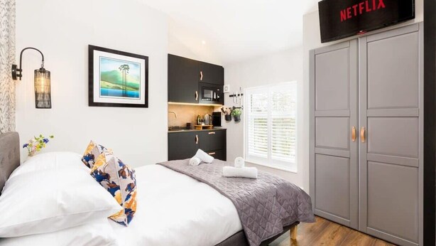 A bedroom with a bed, a wardrobe, a kitchenette, a framed picture, a wall-mounted lamp, and a sign indicating a Netflix service. - Second floor compact studio with mini kitchen (Warrington)