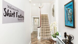 A hallway with a sign reading "Welcome to the Shirt Factory Apartments," a staircase with patterned carpet, and a framed artwork on the wall. - Second floor compact studio with mini kitchen (Warrington)