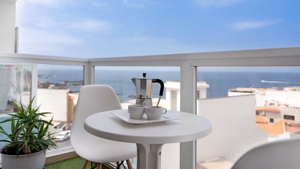 Un balcon avec une table blanche, deux chaises blanches, une plante en pot et une vue sur la mer.