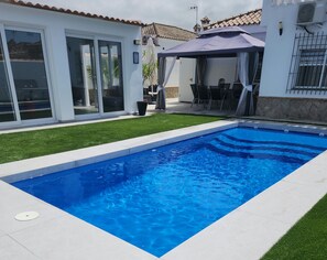 Une piscine avec une terrasse carrelée, un patio couvert et un bâtiment blanc doté de grandes fenêtres.