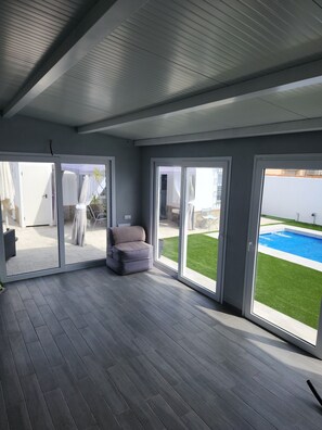 Een ruime kamer met een grijze bank, grote schuifdeuren en uitzicht op een zwembad.