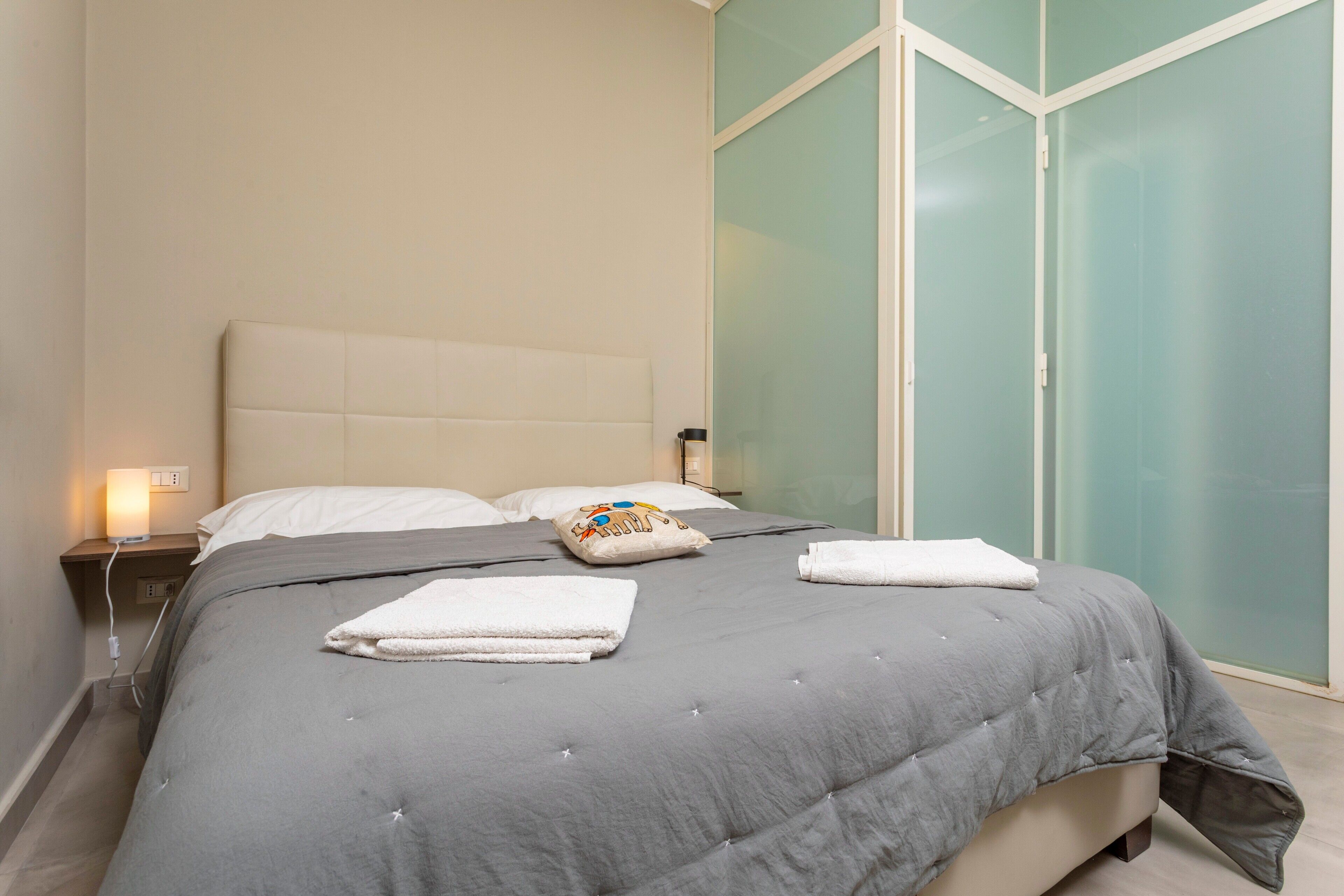 Una cama bien hecha con edredón gris, almohadas blancas y una almohada decorativa. Se observa una mesita de noche con una lámpara y un enchufe.