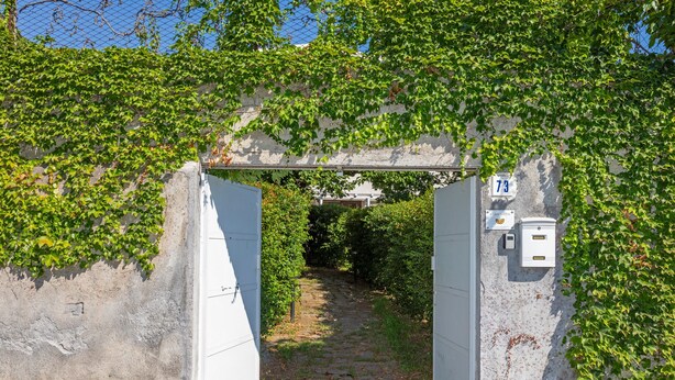 Ein gepflasterter Weg führt durch ein Rundbogenportal mit efeuüberwachsenen Mauern und einem Schild mit der Nummer 73.