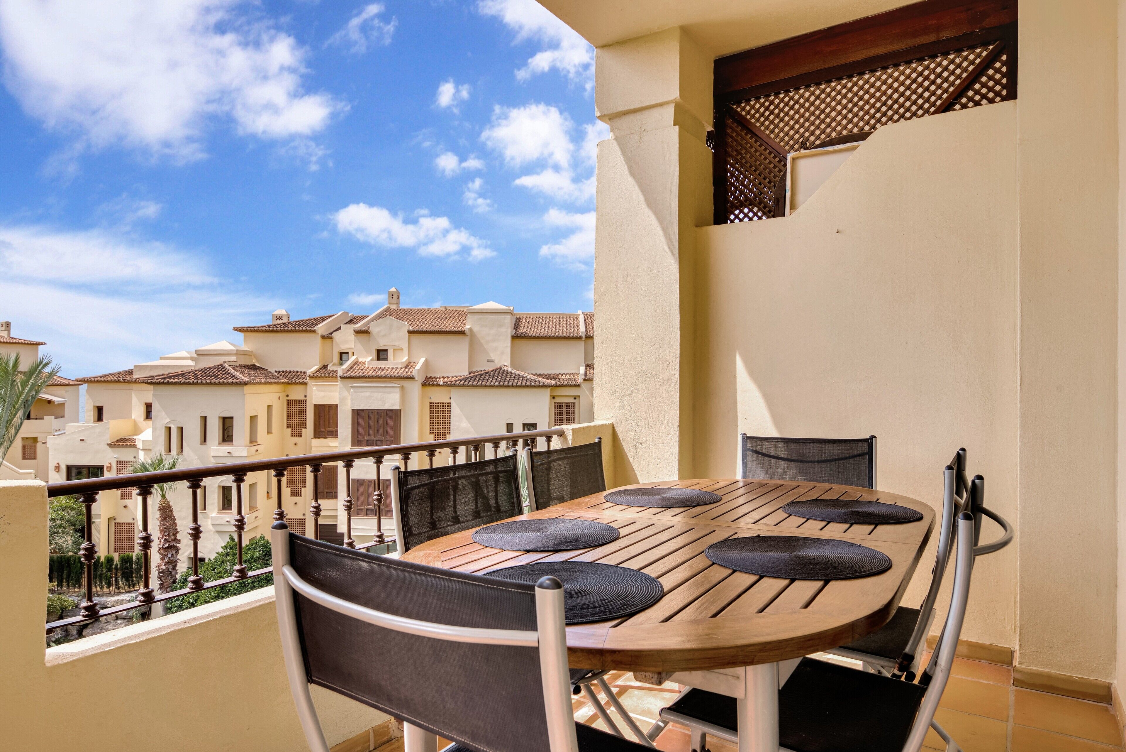 Un balcon avec une table et des chaises en bois, une vue sur les immeubles voisins et un ciel dégagé.