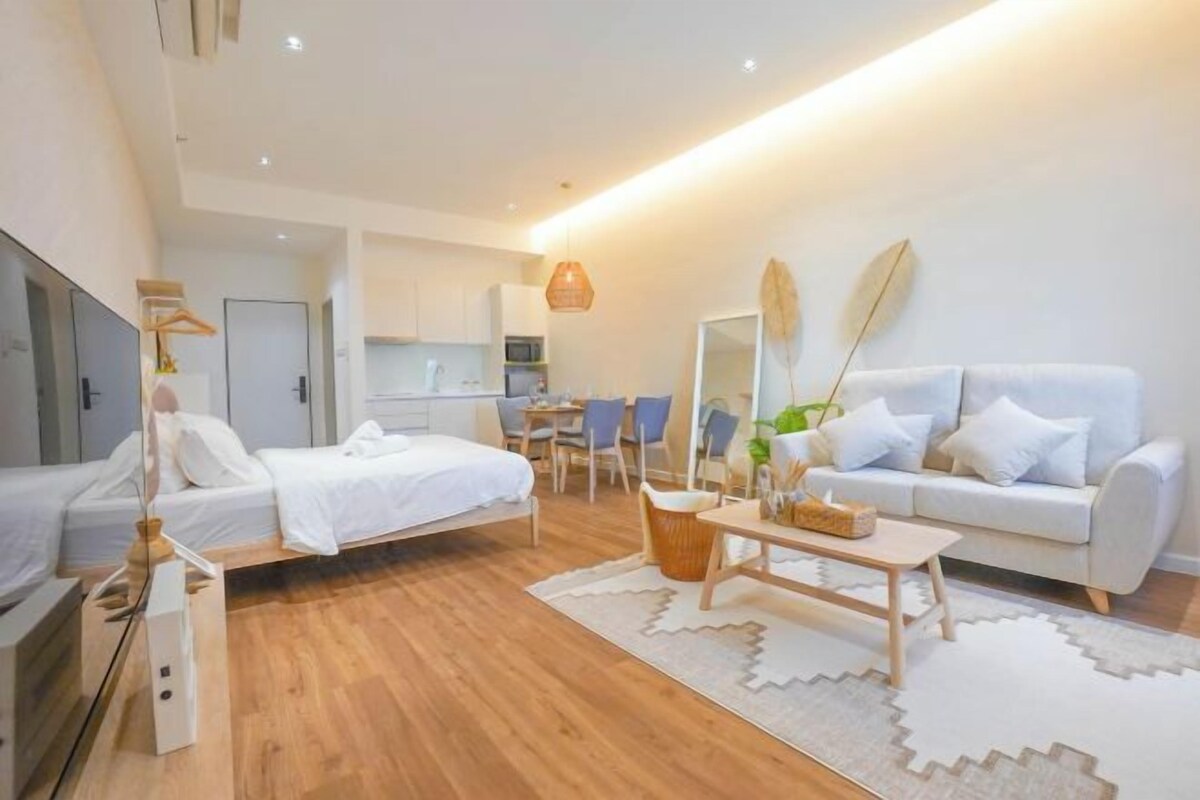 A modern hotel room with a bed, a sofa, a dining area, and a kitchen in the background.