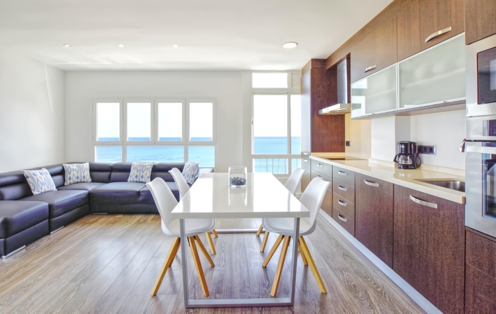 A modern kitchen with a dining area, large windows, and a view of the sea.