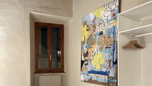 Une chambre à coucher comprenant un lit, une commode, un radiateur, une fenêtre et un mur orné d’un tableau abstrait aux couleurs vives.