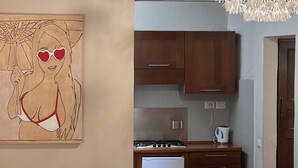 Une cuisine compacte avec des meubles en bois, une cuisinière à gaz et un petit réfrigérateur. On y trouve un coin repas avec une table recouverte d’une nappe à motifs floraux et des chaises blanches. Un luminaire original est visible au-dessus de la table à manger.