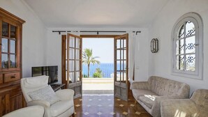 A living room with a TV, two sofas, a cabinet, and a view of the sea through open doors.