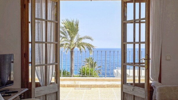 An open door leading to a balcony with a view of the sea and palm trees.