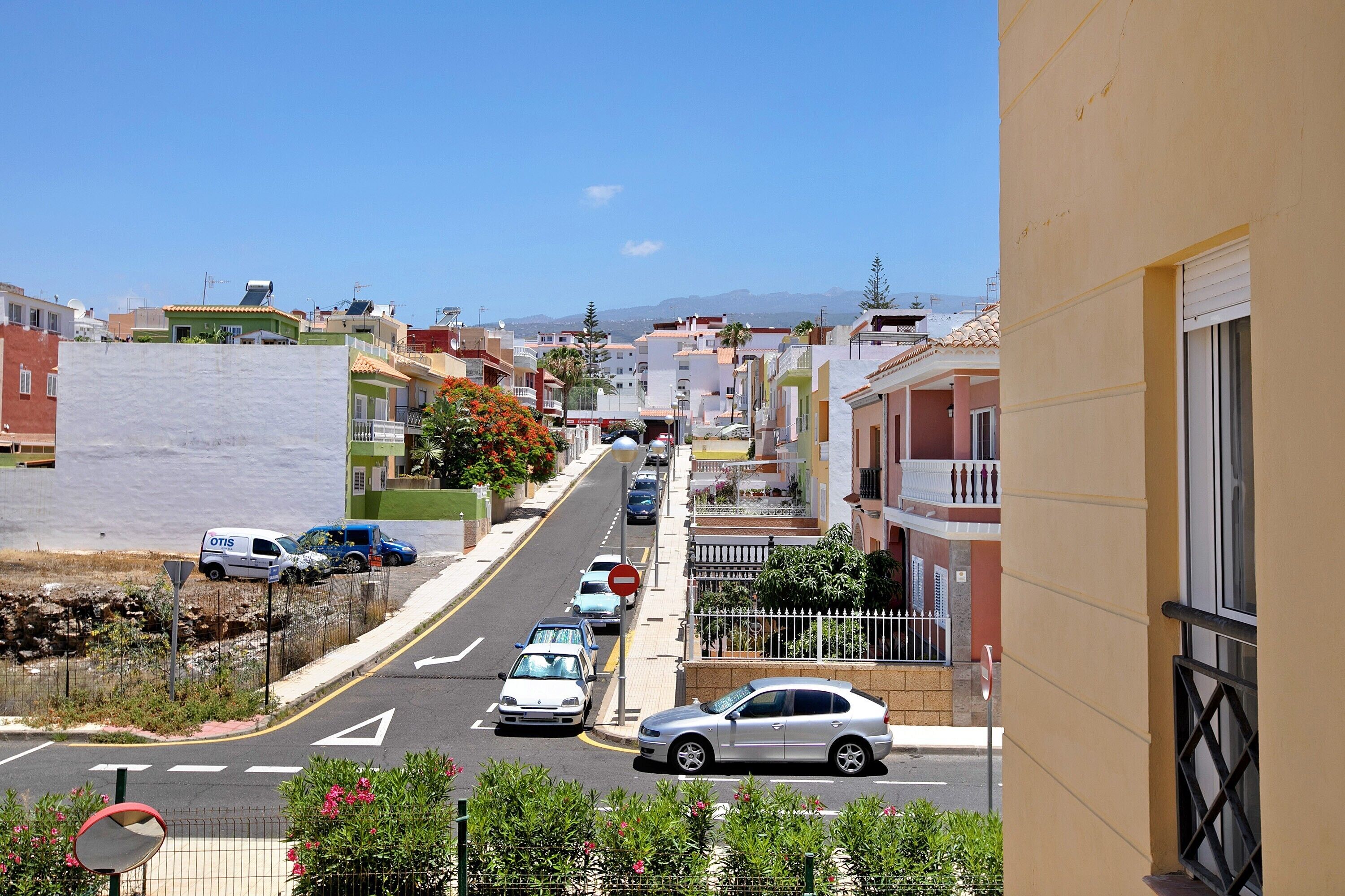 A residential street with parked cars, a clear sky, and buildings with balconies.