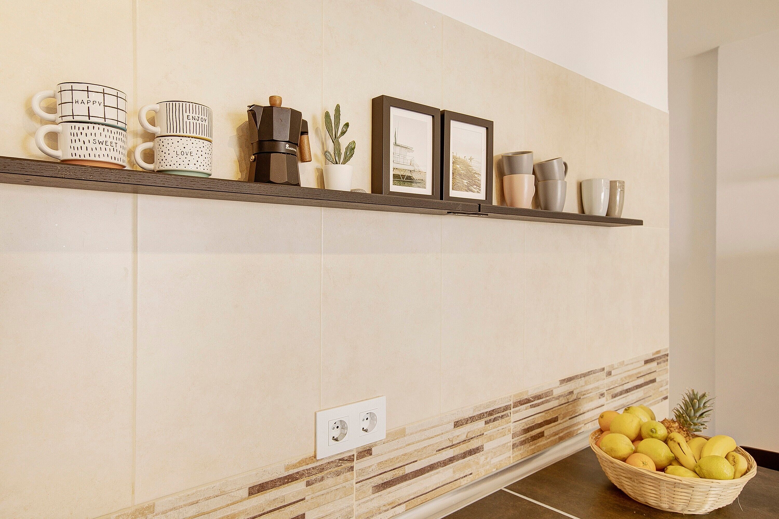 A kitchen with a shelf holding mugs, a coffee maker, a cactus, framed pictures, and a fruit basket.